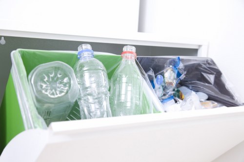 The image shows a close-up view of a partially open white waste collection bin situated inside a clean, modern environment, possibly a kitchen or utility room. Inside the bin, there is a green plastic liner or compartment holding three clear plastic bottles, two with blue and red caps, respectively. The bottles appear to be made of lightweight PET material, with some displaying slight deformation from previous use. To the right of the bottles, there is a black plastic bag filled with miscellaneous waste items, including smaller plastic wrappers and packaging. The bin's interior surface is textured plastic, and the environment around the bin is well-lit with natural or artificial bright light, emphasizing the clarity and transparency of the bottles and the cleanliness of the space. The surrounding background includes a white shelf or countertop edge, suggesting a typical domestic or commercial setting where rubbish is collected prior to removal, possibly following a service from Commercial Waste Removal Highbury. The scene reflects proper waste segregation practices consistent with rubbish removal procedures in London postcodes similar to N5 or N7, in the vicinity of Highbury.