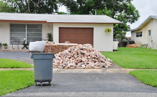 A residential driveway in Highbury is shown with a large mound of mixed light brown and beige bricks stacked directly on the pavement, indicating debris awaiting removal. To the left of the brickpile, there is a dark grey wheeled rubbish bin with a closed lid positioned on the asphalt surface. Behind the pile, a single-story house with white exterior walls and large front windows featuring horizontal blinds is visible, alongside a closed, brown sectional garage door. The house has a paved area and a small grassy patch with well-maintained green lawn surrounding the construction debris. To the right, part of another similar residence is visible, with a glimpse of its garden and outdoor items. The scene suggests a domestic residence undergoing renovation or clearance, and the image is associated with services such as those provided by Commercial Waste Removal Highbury that specialise in rubbish disposal. The overall lighting indicates daytime with overcast skies, highlighting the details of the debris, environment, and surroundings clearly.