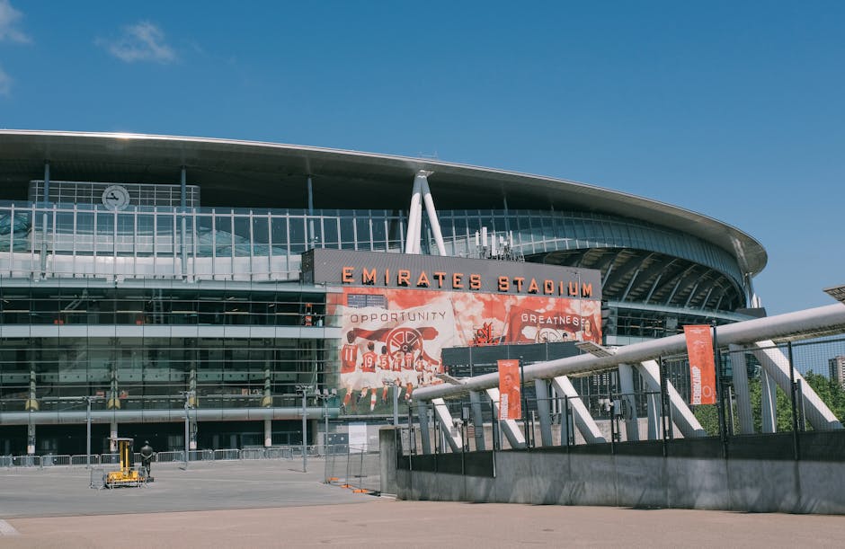 An empty view of the interior of a football stadium with a focus on the tiered seating sections, which are predominantly red with some grey and white areas, and the green pitch visible at the bottom o