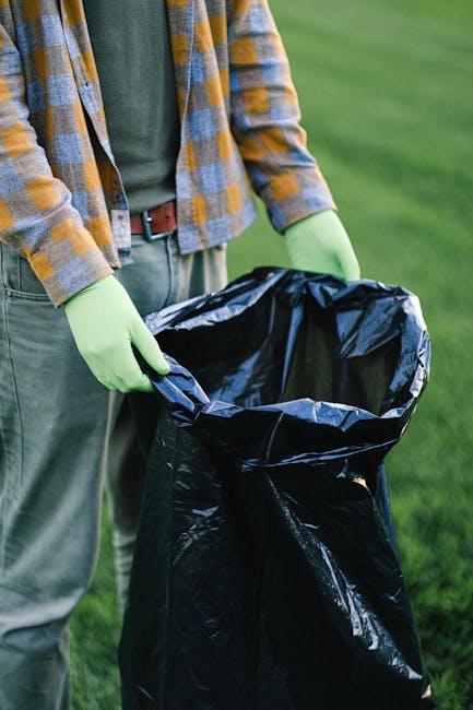 Three large black plastic rubbish bags, some with visible printed labels, are piled against a dark metal fence situated on a concrete pavement. The bags appear to contain waste materials and are place
